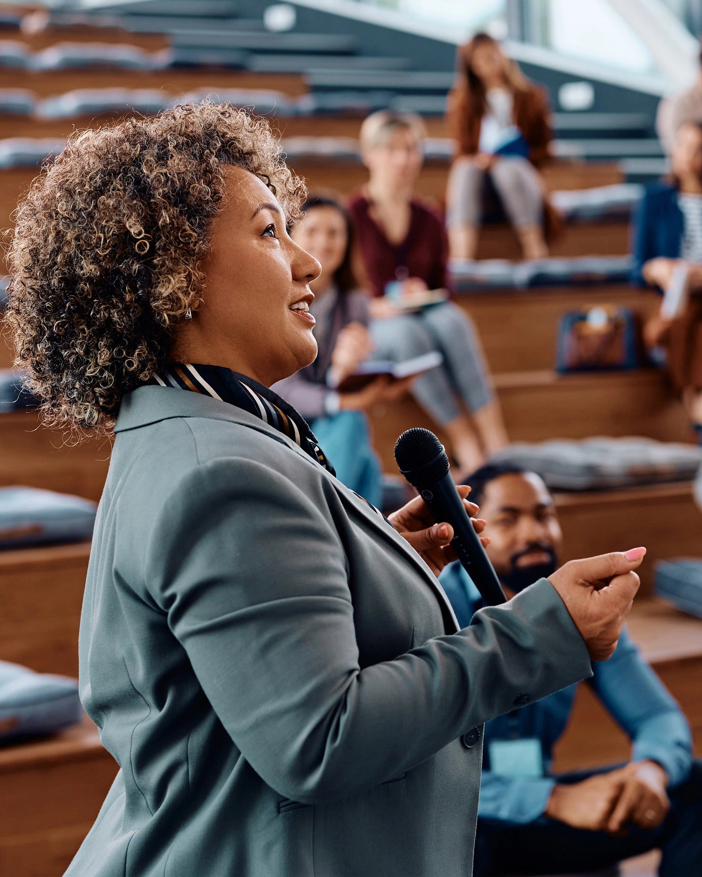 Woman speaking into a microphone in an auditorium with audience members.