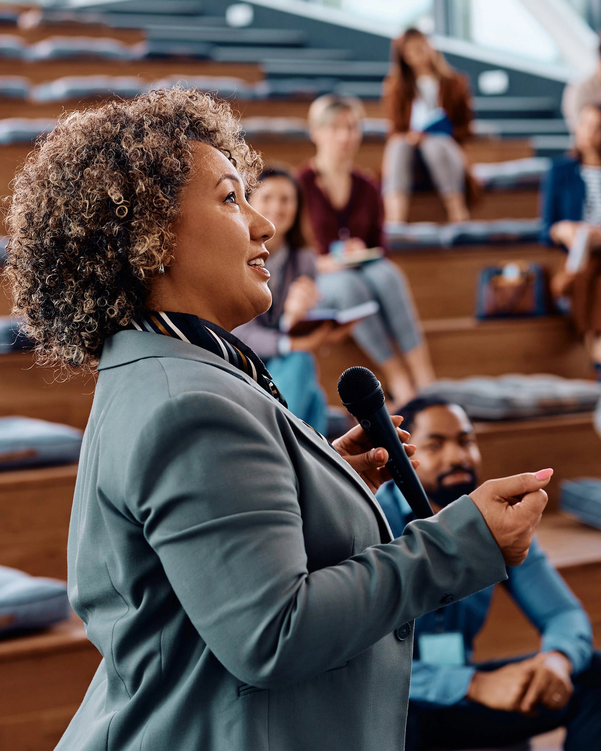 Woman speaking into a microphone in an auditorium with audience members.
