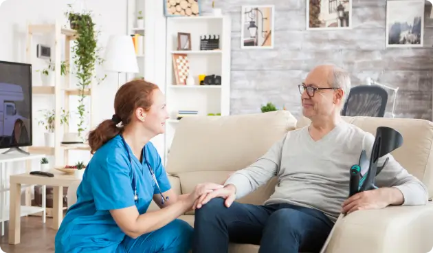 Nurse in blue scrubs sitting on a couch with an elderly man in a living room setting.
